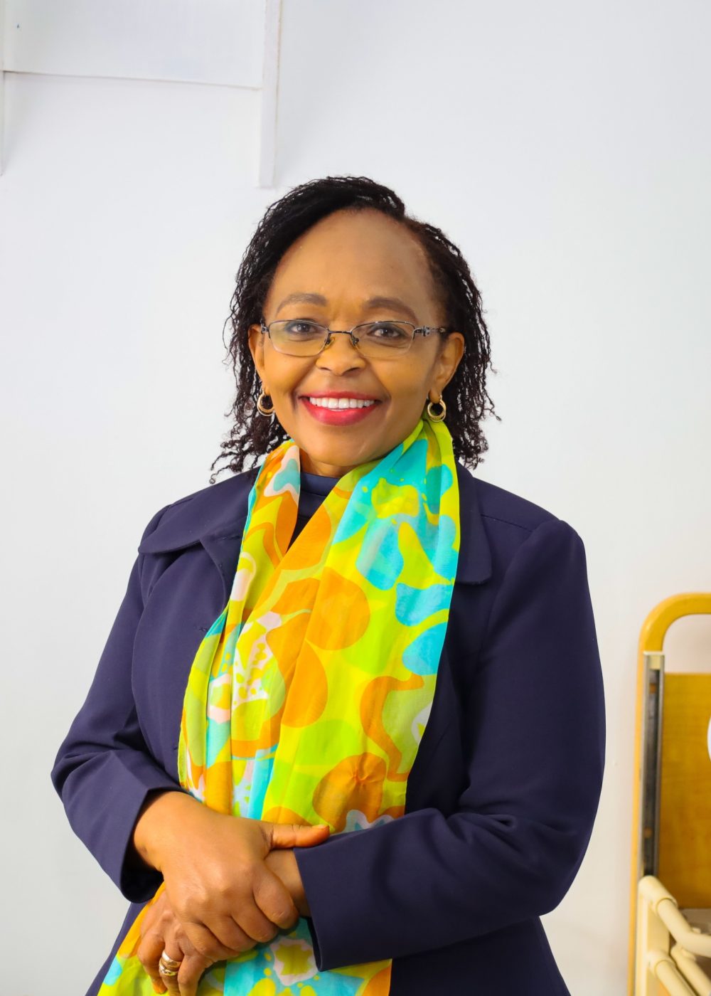 Professional portrait of Faith Malavu smiling confidently in formal attire, standing in a modern healthcare or office setting, representing leadership, compassion, and professionalism in healthcare services.